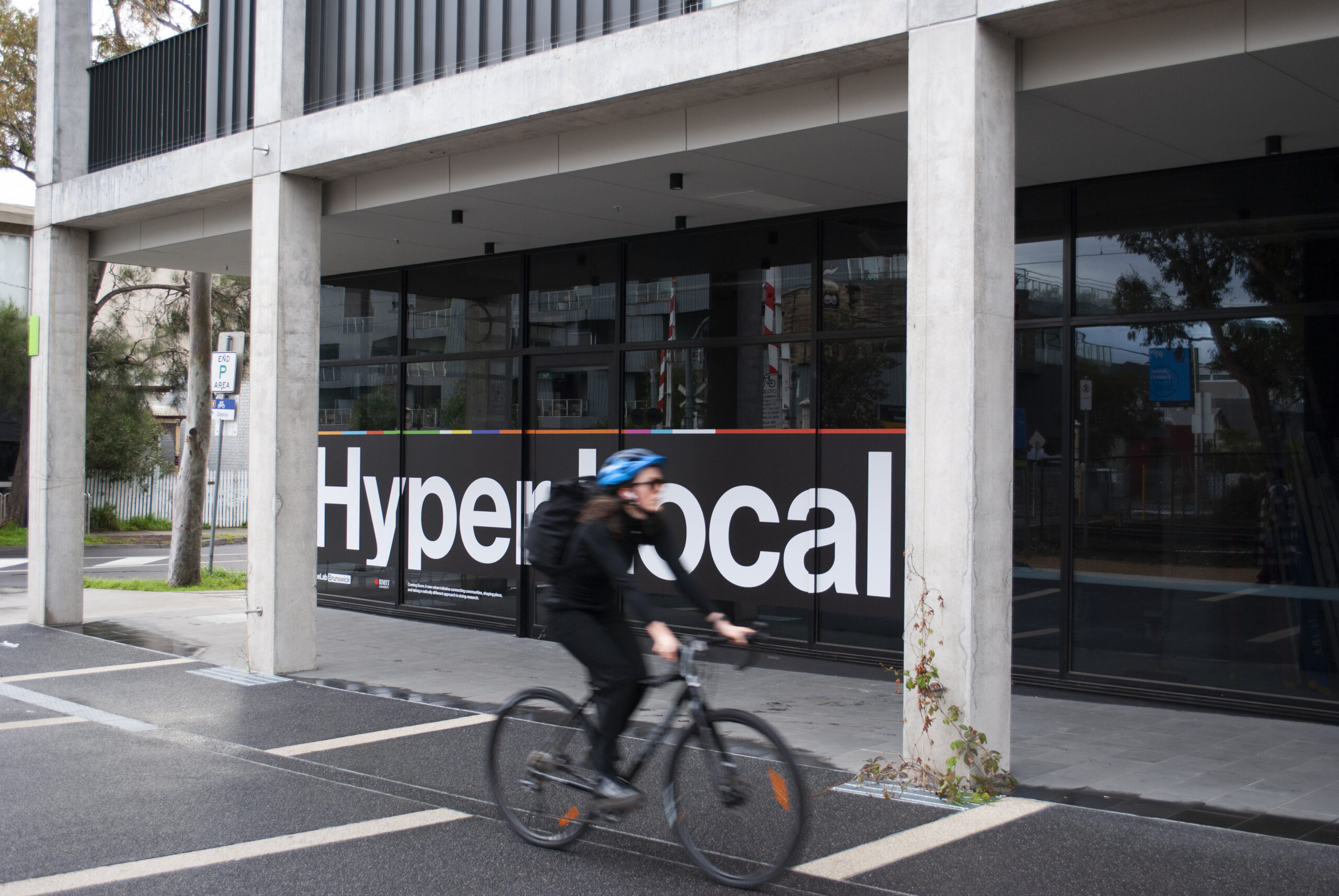 A cyclist riding past the PlaceLab Research Studio. A sticker on the glass window reads Hyperlocal.