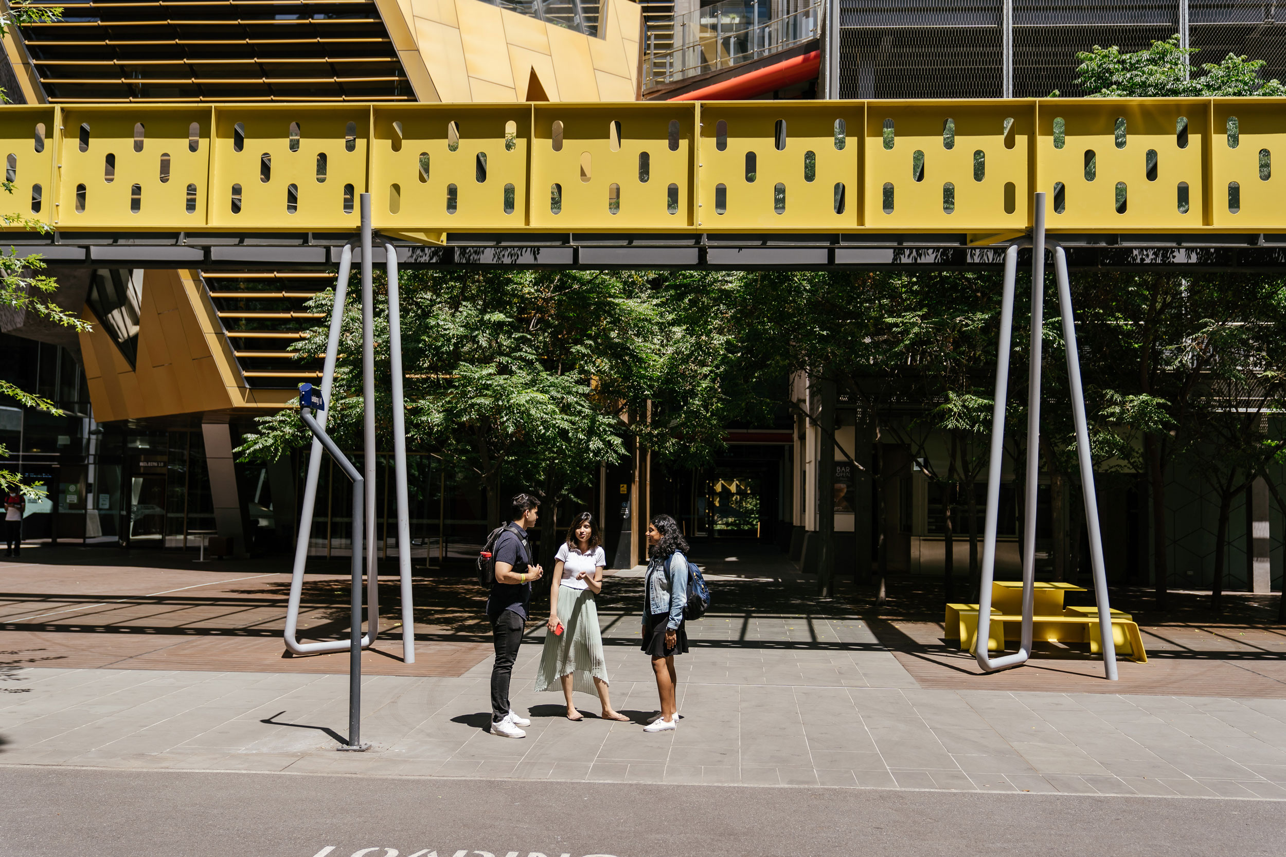 A group of three students standing beneath a yellow architectural structure.